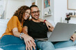 © Jake Jakab/ADDICTIVE STOCK - Cheerful young diverse couple in casual clothes and eyeglasses holding hands while sitting together on sofa and having video call on laptop