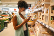 © Jake Jakab/ADDICTIVE STOCK - Modern young black woman in protective mask using app on mobile phone for checking information about product while making purchases in supermarket