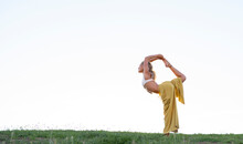 Side View Full Body Barefoot Female In Bra And Loose Trousers Performing Dancers Pose And Outstretching Hand With Cup Of Coffee While Practicing Yoga On Grassy Hill Against Urban Environment