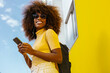 © Victor Torres/ADDICTIVE STOCK - Black woman with afro hair listening to music on mobile in front of a yellow wall