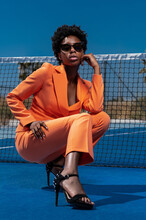Young Stylish Black Woman In Orange Apparel With Sunglasses And High Heeled Sandals Squatting Near Fence On Sports Ground Looking At Camera