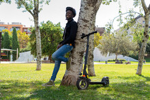 Side View Of Cheerful African American Male Wearing Helmet Leaning On Tree In Urban Garden And Standing Near Electric Scooter While Looking At Camera