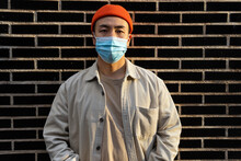 Concentrated Asian Male In Orange Bennie And Protective Face Mask Standing Calmly On Brick Wall Looking At Camera