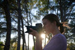 © German Linares/ADDICTIVE STOCK - Side view of young female photographer with professional photo camera shooting pictures while standing in green forest in sunny day