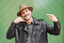 Trendy Friendly Male In Hat And Leather Jacket Smiling And Looking At Camera Doing Call Me Gesture With Hands On Green Gate Background