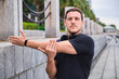 © Luis Gomez/ADDICTIVE STOCK - Concentrated male athlete in sportswear and headphones standing on street and warming up arms before workout in city