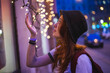 © Nacho Zaitsev/ADDICTIVE STOCK - Side view of stylish hipster millennial woman in hat and with backpack touching glowing garland while standing near building in evening city