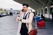 © Manuel Ruiz Alba/ADDICTIVE STOCK - Busy man with dark hair in suit and coat speaking on phone and looking at watch while standing near airport terminal at daytime
