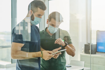 Through glass busy male programmers in medical masks standing with tablet in office and discussing software development at work during coronavirus outbreak