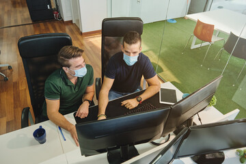From above of male coworkers in medical masks sitting together using program for software during coronavirus pandemic