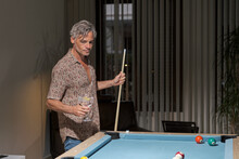 Thoughtful Mature Man Enjoying Cocktail While Holding Cue Stick An Playing Billiard In Bright Room