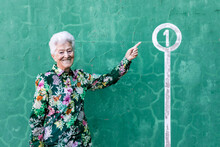 Happy Aged Gray Haired Female In Colorful Blouse Standing Against Green Wall And Pointing At Number 1 Sign On Parking Lot