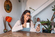 © Txema Gerardo/ADDICTIVE STOCK - Content adult ethnic female in warm knitted wear sitting in chair and messaging on mobile phone while resting with cup of hot drink in cozy house