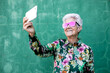 © Sergio Victor Vega/ADDICTIVE STOCK - Happy aged gray haired female in stylish outfit and sunglasses taking selfie on mobile phone while standing against green wall