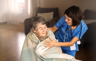 A young nurse takes care of an elderly woman at an 80-year-old home, wraps a blanket around her. Happy retired woman and trust between doctor and patient. Medicine and healthcare.