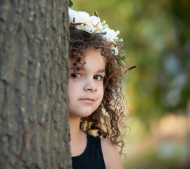 Wall Mural - Curly haired mixed race girl in floral wreath peeking from behind tree