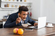 african american freelancer in suit using laptop while eating breakfast near gadgets.