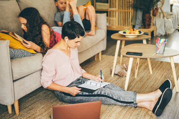 Short haired woman with bluetooth earpiece writes in notebook sitting on floor near laptop while teenage girl and boy lies on sofa in light living room.