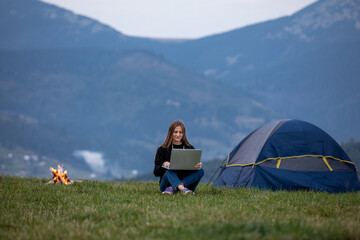 young female freelancer working on laptop in the mountains in the evening. Tourist girl sitting near campfire and tent. Copy space