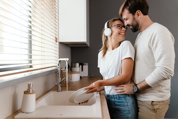 Wall Mural - Beautiful cheerful couple smiling and hugging while washing dishes