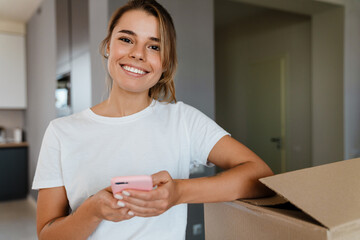 Wall Mural - Beautiful girl holding cellphone by cardboard boxes in new apartment