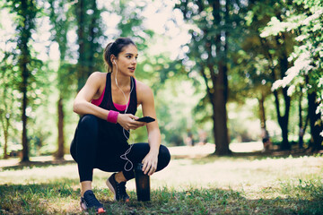 Young attractive fit caucasian sportswoman crouching in nature and using smart phone to search music for running.