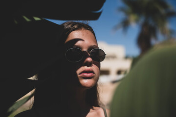 Wall Mural - cute young teenage girl or woman posing with banana tree leaves dressed in summer hipster vintage white stylish clothes. Trendy girl posing. Funny and positive woman in sunglasses