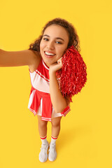 Wall Mural - Beautiful young cheerleader taking selfie on color background