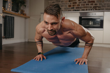 Wall Mural - A muscular man with a beard is doing pushups on a blue yoga mat in his apartment in the evening. An athletic guy with tattoos on his forearms is doing a chest and triceps workout at home.