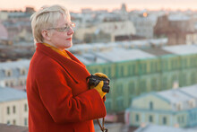 A Mature Woman In Glasses With Short Blond Hair In Red Coat Stands With Binoculars On The Roof Of An Old House In St. Petersburg