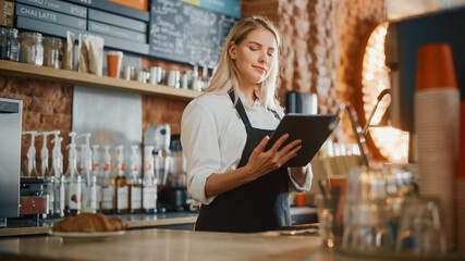 Beautiful Young Caucasian Coffee Shop Manager with Blond Hair is Making Notes on a Tablet Computer and Orders Inventory Items for the Menu in a Cozy Loft-Style Cafe. Successful Restaurant Owner.