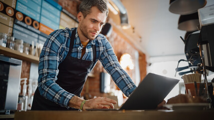 Young and Handsome Coffee Shop Owner is Working on Laptop Computer and Checking Inventory in a Cozy Cafe. Happy Restaurant Manager in Checkered Shirt Browsing Internet and Chatting with Friends.