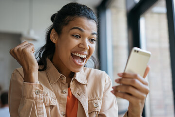 Happy emotional girl celebrates victory, sports betting concept. Young Excited African American woman using mobile app for online shopping with cash back
