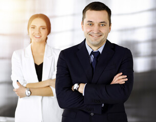 Portrait of a friendly middle aged businessman in a dark blue suit, standing with crossed arms together with a colleague in a sunny modern office. Successful business people concept