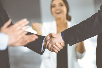 Businessmen are shaking hands while standing with colleagues applauding in office, close-up. Business success concept