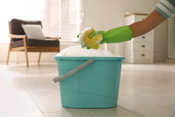 Poster - Woman holding sponge with foam over bucket indoors, closeup. Cleaning supplies