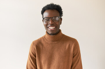 Wall Mural - Nerdy young African guy in glasses and being sweater smiling broadly at camera, expressing positive emotions, rejoicing at good news. Human facial expressions, feelings, reaction and life perception