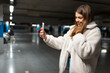 © Andrii  - Beautiful girl takes selfie in the underground parking. Fasionable young woman with smartphone