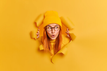 Beautiful surprised redhead beautiful woman has wondered face expression opens mouth stares at something unbelievable looks through hole in yellow paper background wears hat transparent glasses