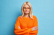 © wayhome.studio  - Portrait of beautiful serious middle aged woman keeps hands crossed wears spectacles and orange sweater looks confidently at camera listens attentively interlocutor isolated over blue background