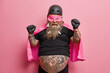 © wayhome.studio  - Positive male superhero raises clenched fists celebrates something with triumph has thick beard and tattooed belly wears helmet gloves and cape isolated over pink background. Powerful successful man