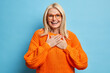 Portrait of blonde female with cheerful expression keeps hands pressed to chest expresses gratitude for heartwarming compliment wears eyeglasses and orange sweater isolated over blue background