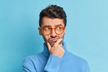 Wall Mural - Headshot of displeased pensive adult man holds chin purses lips and looks aside tries to make decision wears spectacles and turtleneck isolated over blue background. People and thoughts concept