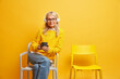 © WHstudio Leushin N - Studio shot of middle aged woman sits on comfortable chair wears transparent glasses holds mug of coffee listens favorite music via stereo headphones isolated over yellow background. Lifestyle concept