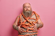 © wayhome.studio  - Puzzled bearded man wrapped with sausages has fat tattooed belly excess weight isolated over pink background. Obese adult male human eats junk food. Harmful nutrition and unhealthy lifestyle