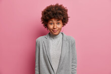 Satisfied African American Woman In Grey Formal Clothes Feels Happy After Successful Working Day Has Curly Bushy Hair Isolated On Pink Background. Glad Female Teacher Listens Attentively Pupils Answer