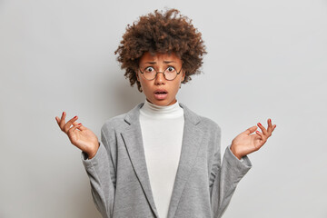 Poster - Puzzled dissatisfied young woman with curly Afro hair spreads palms sideways wears round spectacles grey formal clothes poses indoor. Indignant businesswoman cannot understand whats problem.