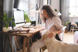 © AnnaStills - Young woman sitting at the table in front of computer at home and feeding her dog