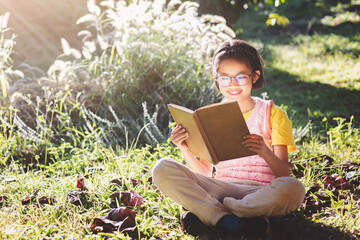 Canvas Print - cute little girl sitting on the grass reading a book in garden at summer time