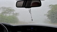 Driving a car on the road on a rainy day, wiper blades cleaning the windshield quickly. Praying rosary with a cross hanging in the rearview mirror.